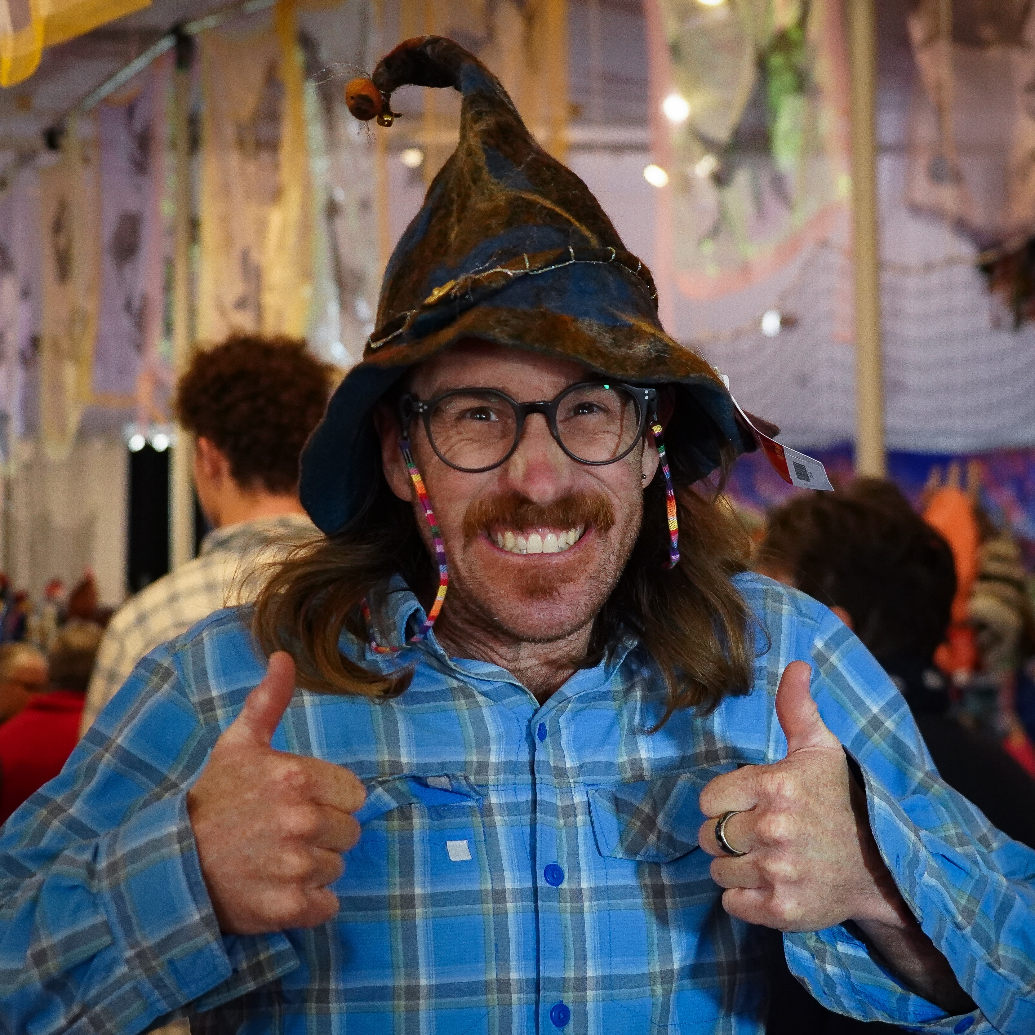 A man giving two thumbs up as he smiles at the camera, he is wearing a pointing handmade hat on his head.