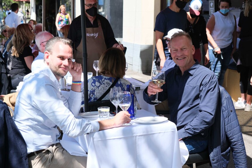 Two men sitting at an outdoor restaurant as one raises his glass in a toast.