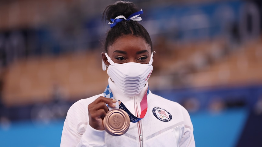 Simone Biles wears a mask but appears to be smiling as she holds up her bronze medal.