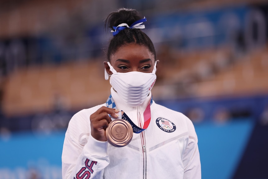 Simone Biles wears a mask but appears to be smiling as she holds up her bronze medal.