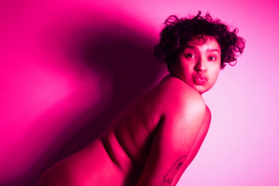 A nude person with curly hair faces side on and makes a kiss face while bathed in pink lighting