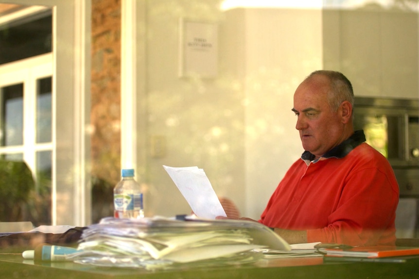 A man is seen through a window looking at a document. There are a pile of papers in front of him.