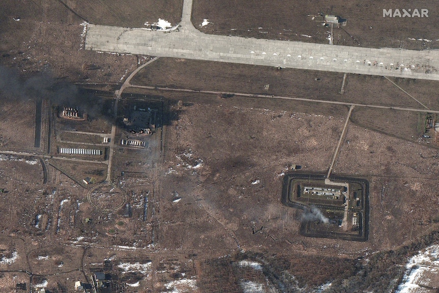 Runway of airport near top of frame, black smoke coming from burning fuel depot, white smoke from burning building