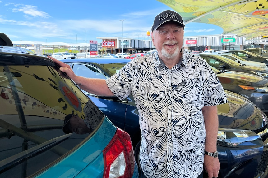 A man stands beside his car in the supermarket carpark.