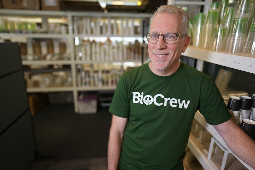 A middle-aged white man with glasses and a 'BioCrew' shirt standing in front of shelves containing takeaway containers and cups