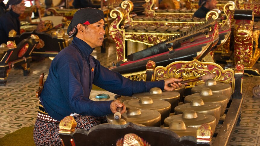 A close up of a Javanese bonang player sitting on the ground, surrounded by other gamelan players.