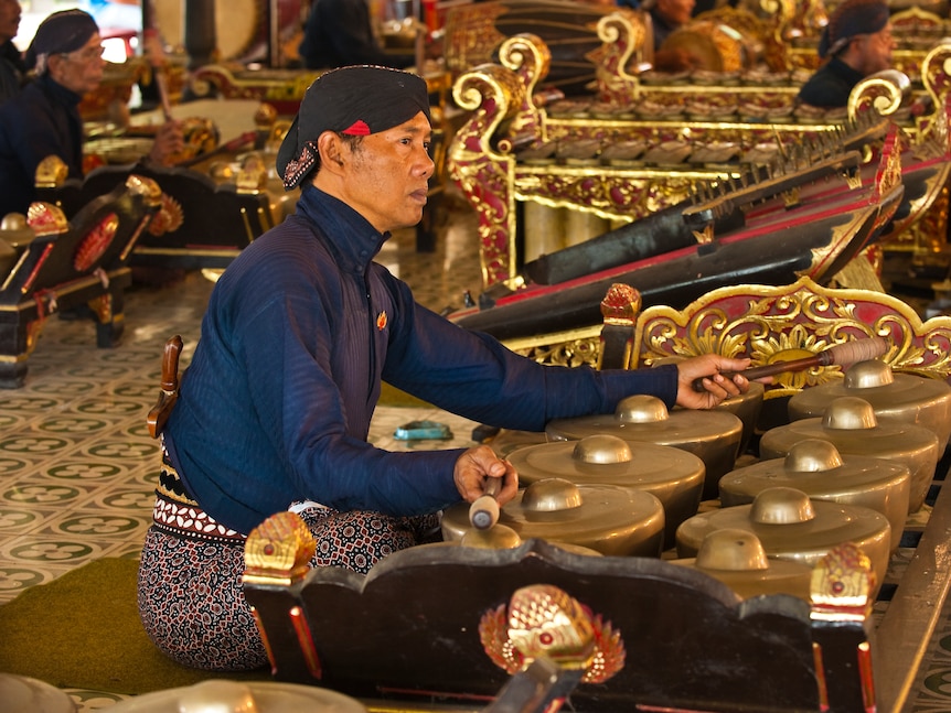 A close up of a Javanese bonang player sitting on the ground, surrounded by other gamelan players.