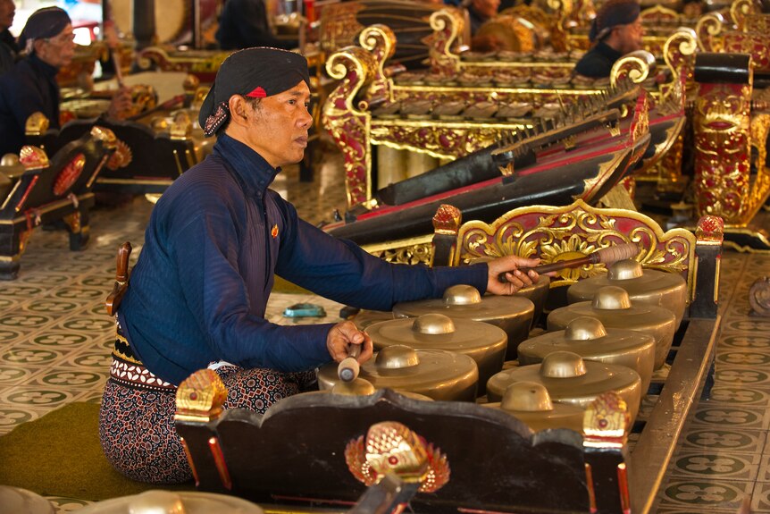 A close up of a Javanese bonang player sitting on the ground, surrounded by other gamelan players. 