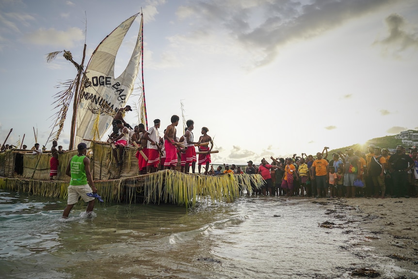 A traditional sailing ship on the shore of a beach surrounded by PNG locals.
