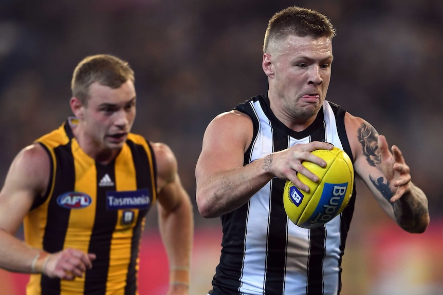 A male AFL player holds a yellow football in his right hand as he is chased by an opposition player.