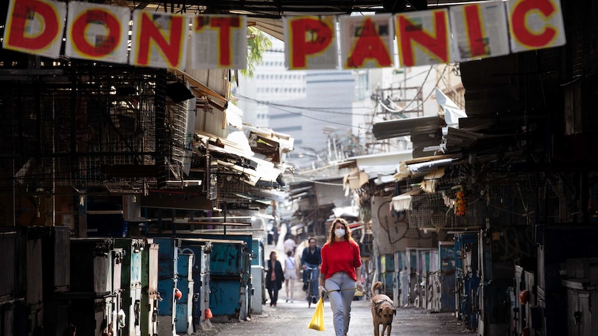 A woman in a face mask walks her dog through Tel Aviv under a sign reading "don't panic"