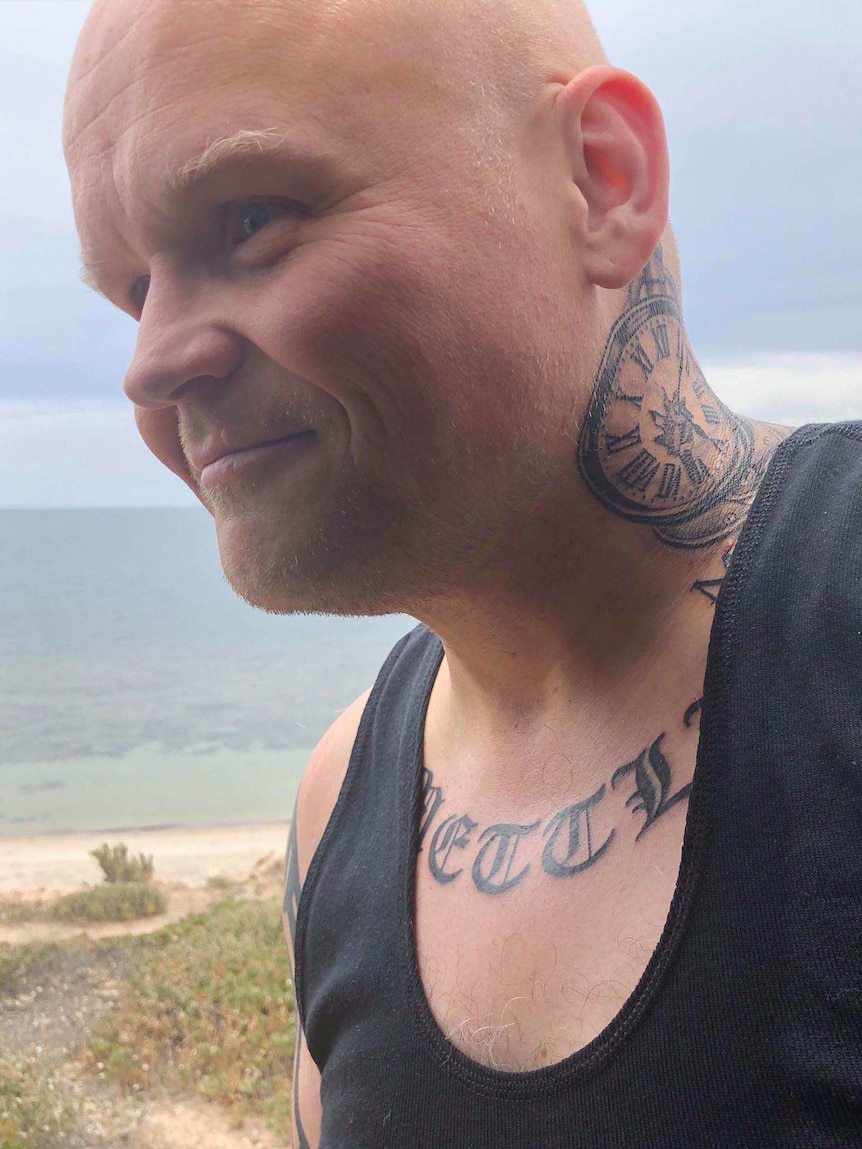 A bald man with tattoos walking along a beach.