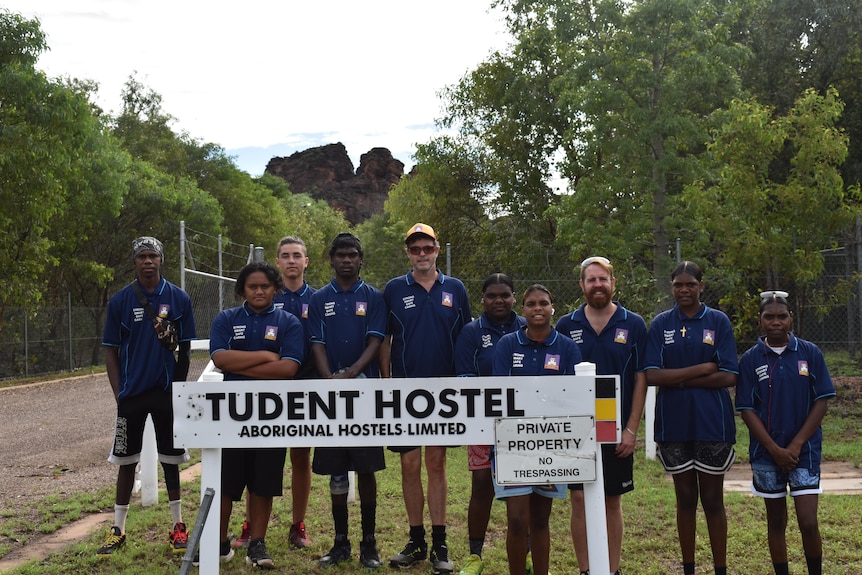 Mostly Indigenous students stand at a hostel sign with trees and a fence in the background