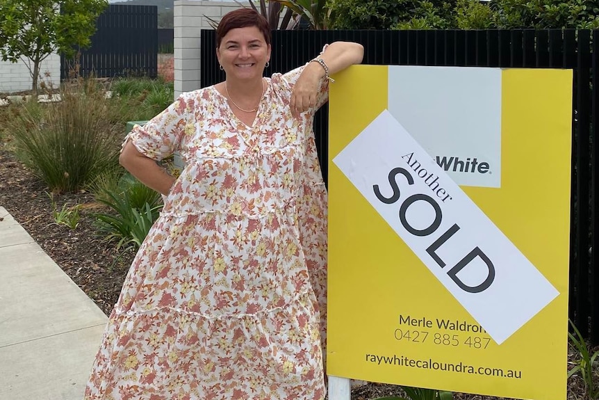 woman stands beside a sold housing sign, smiling