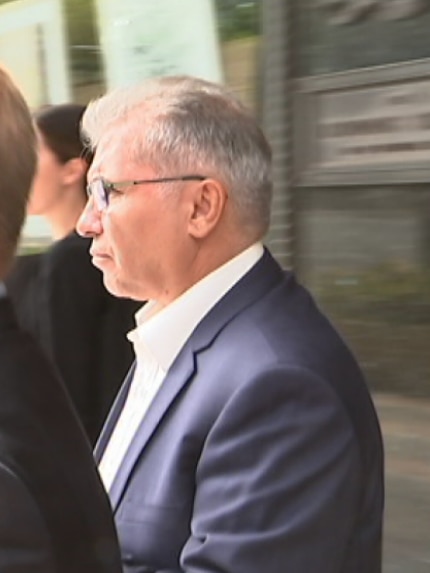 A man wearing a suit and classes, in side profile, walks on a busy street. He is with another man and a woman