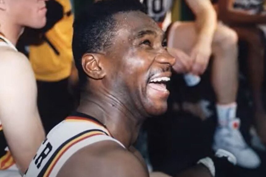 A man with a big smile sits with the rest of his basketball team. 