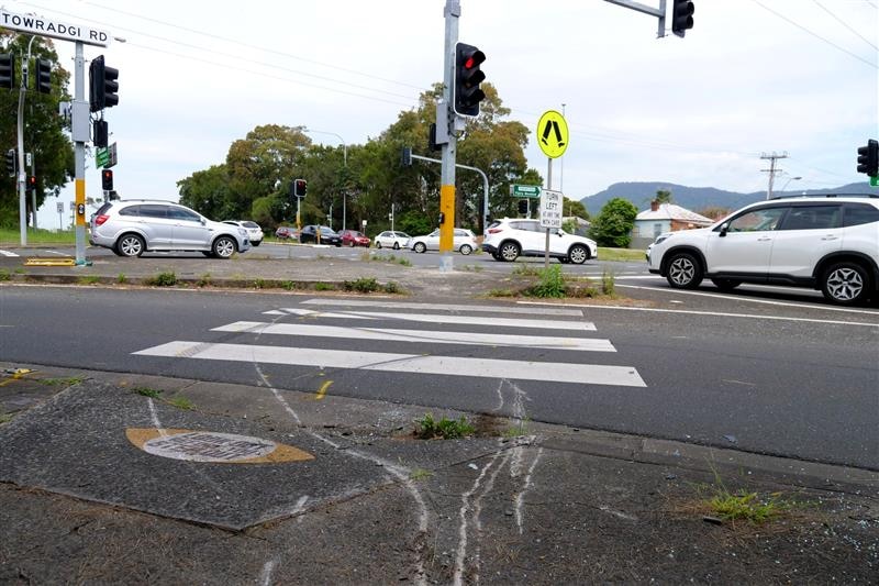 A road and zebra crossing, with surface damage visible on the footpath in the foreground.