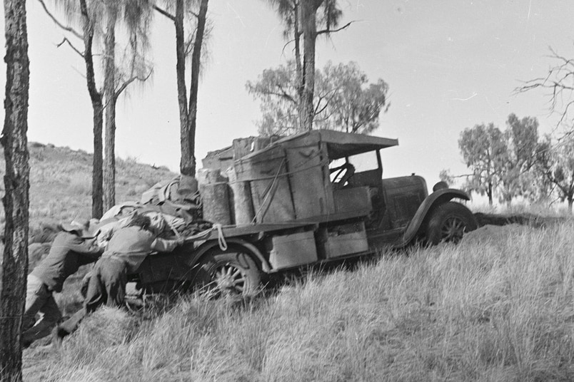 Black and white photo of man pushing cart up a grassy hill.