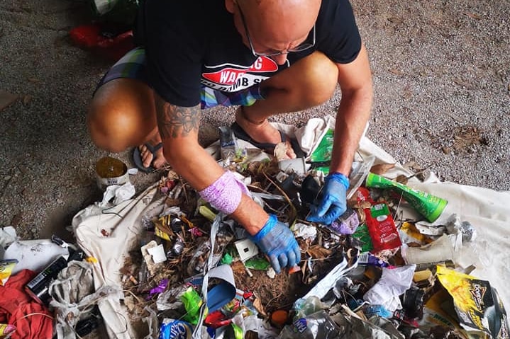 A man wearing blue latex gloves squats on the ground, sorting through a pile of rubbish.