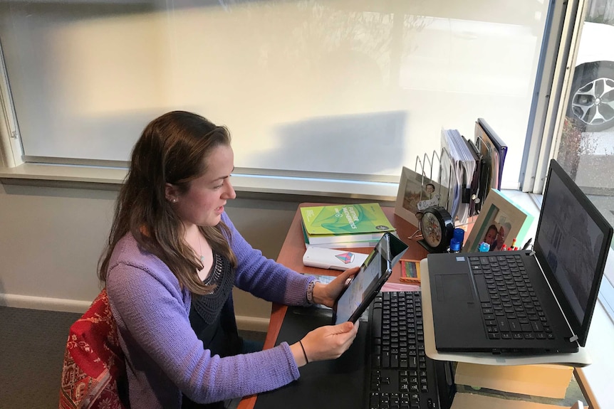 Teacher Claire Glanville sitting at a desk with a PC on it and looking at a separate tablet