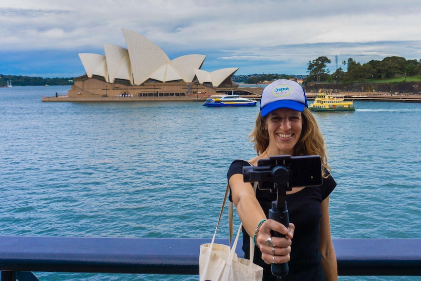 A woman stands on a bridge in front of the opera house with selfie stick