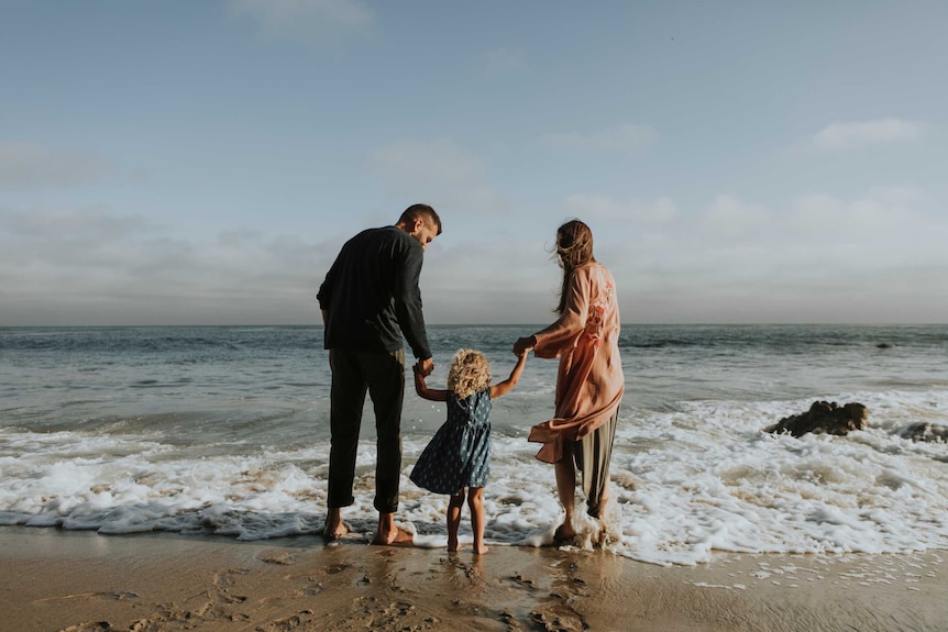 A man and a woman each hold a child's arms while facing the ocean on a beach.