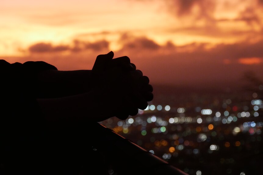 A silouette of two hands over city lights at sunset.