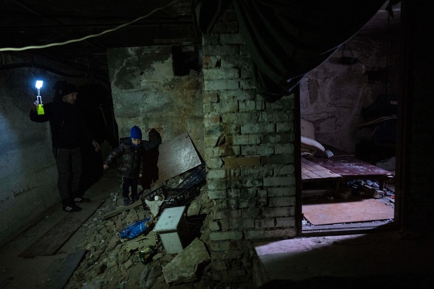A man holds a torch as he walks with his child in a dark basement.