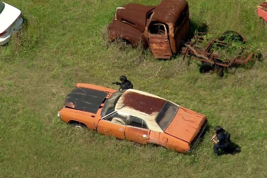 Two police officers with guns crouch next to an old car body on a rural property wearing helmets.