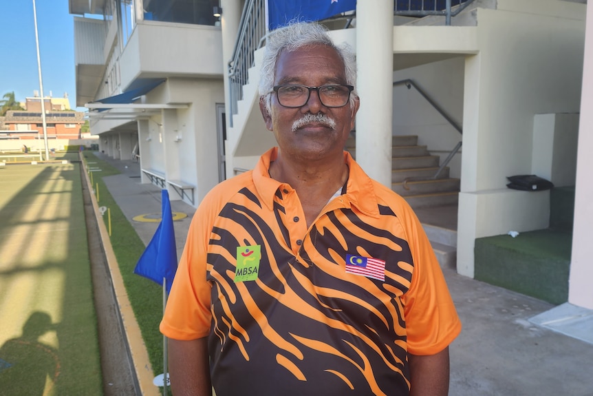 A man with white hair and a moustache stands near a bowling green, wearing a Malaysian sports shirt and smiling at the camera.