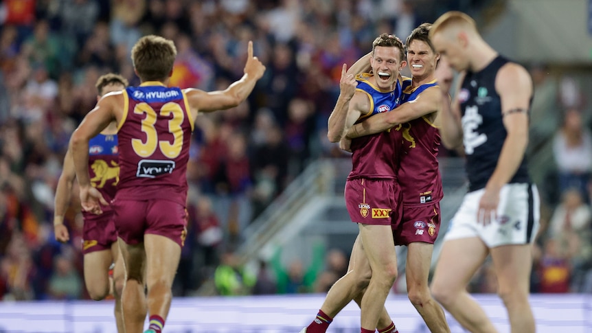 A group of Brisbane players smile and celebrate a goal, as a teammate runs towards them pointing his finger in the air.