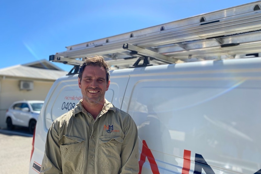 A man with short hair wears a cargo shirt in front of a van and smiles at the camera