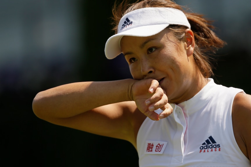 Peng shuai wearing white tennis gear and hat brings her right arm to her face on court