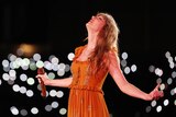 Taylor Swift leans back and closes her eyes on stage in an orange dress as lights blur around her