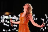 Taylor Swift leans back and closes her eyes on stage in an orange dress as lights blur around her