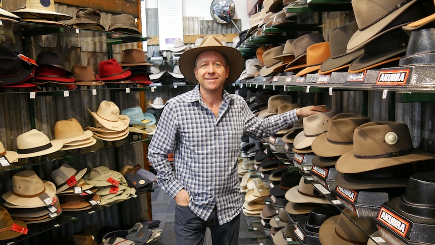 man wearing checked shirt and akubra hat standing in between two shop walls of akubra hats