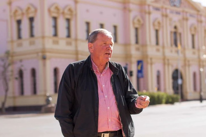 A balding man in foreground in pink shirt in front of town hall.