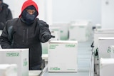 A person in warm cloths and face covering guides a box of vaccines on a conveyor belt.