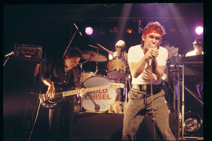 Cold Chisel band performs, circa 1980s. Singer Jimmy Barnes right of frame, in focus, holds mic while wearing red bandana.