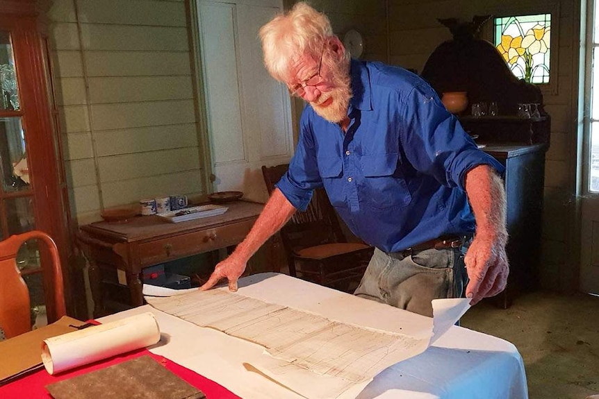 Graham Moffatt stands in his house at Camoola Park station, looking over the long term river height chart rolled out on a table.