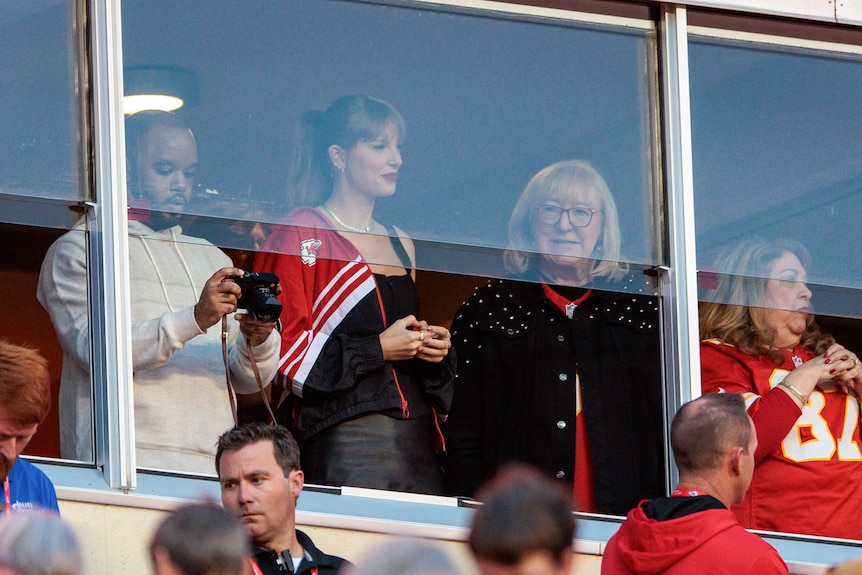 Taylor Swift wearing a jersey stands behind a window with other footabll fans.