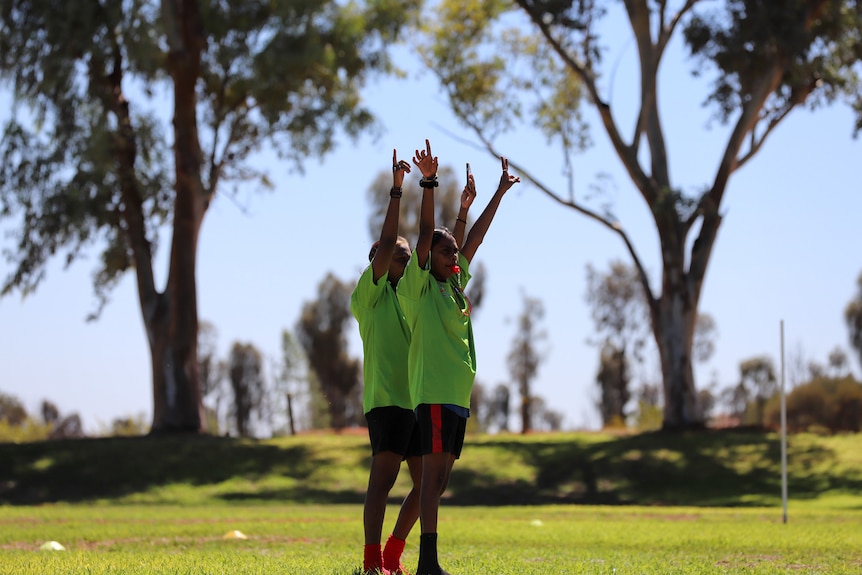 Two young Aboriginal women in lime green umpiring uniforms raise their hands in unison above their heads on a football ground