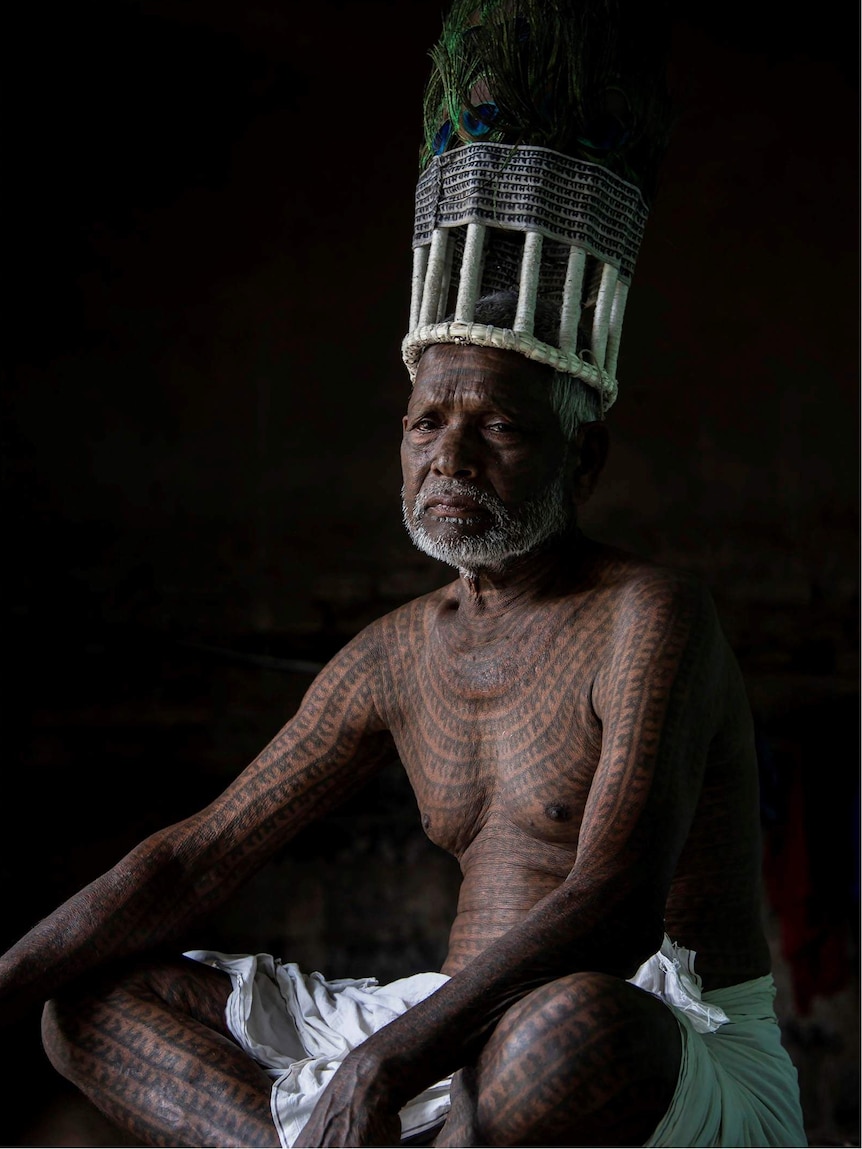 Seated Indian man covered in tattoos wearing a headpiece.