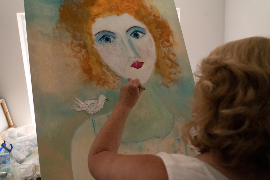 A blonde woman pictured from behind painting a canvas of an auburn haired woman's face.