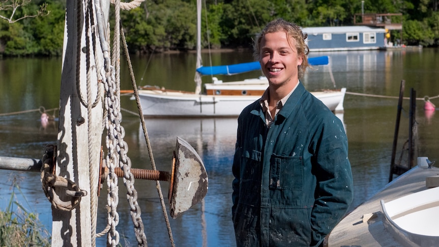 A young man in blue overalls stands on the Brisbane River, besides the boat he built.