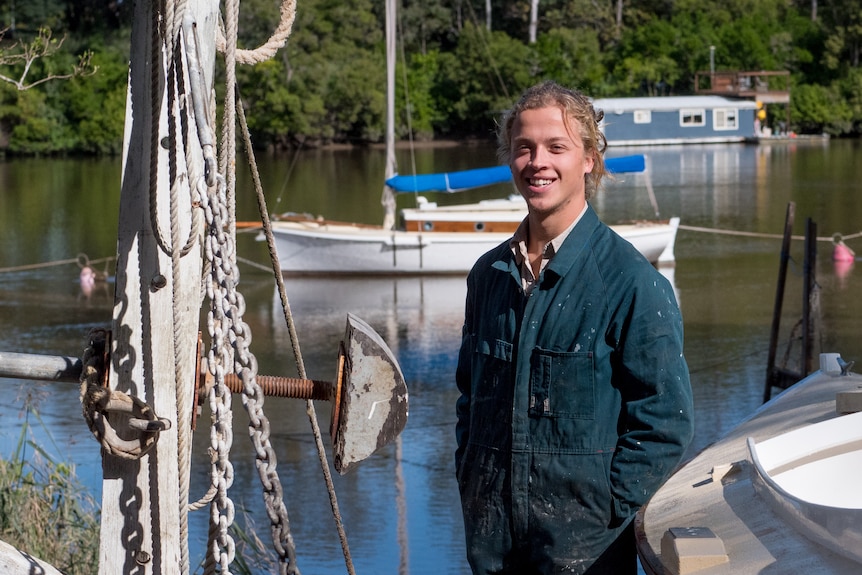 A young man in blue overalls stands on the Brisbane River, besides the boat he built.