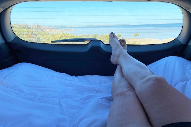Picture taken inside the back of car, with legs in the foreground. Outside the window you can see a wonderful beach view