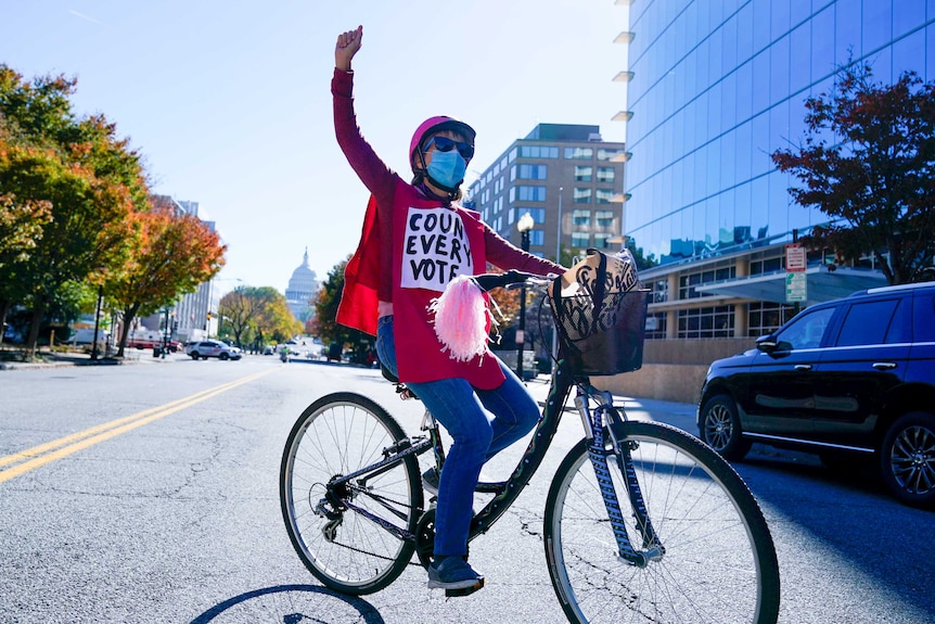 A woman on a bike with her fist in the air and a sign on her chest reading 'count every vote'