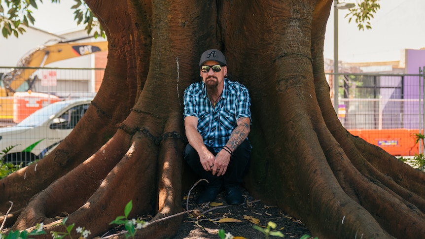 A man in a short-sleeved shirt and baseball cap sits with his back against a tree trunk.