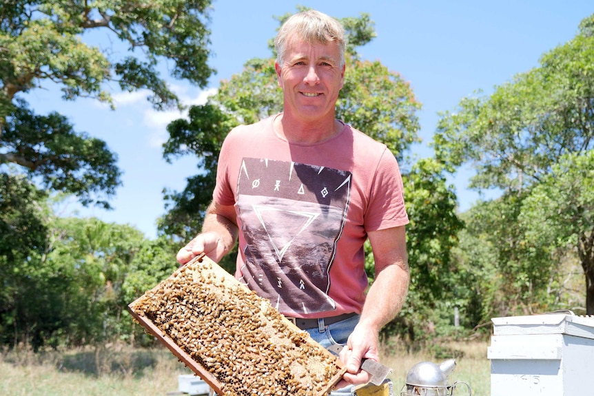 Paul holds a tray from a beehive in front of him, he's smiling at the camera in an open field.